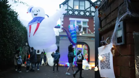 A QR code stuck to the wall outside the house with artificial cobwebs around it. The Halloween house can be seen in the background with people admiring the display.