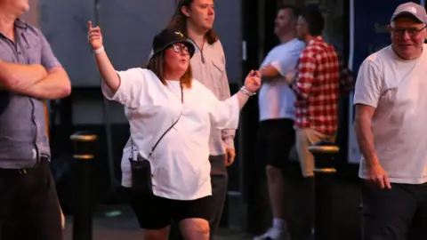 Emma Gibbs A woman in a black bucket hat and a white T-shirt holds her arms wide as she sings in the street surrounded by other people after Oasis at Heaton Park