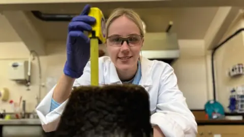 Ellie is looking at the camera and is wearing protective glasses. She is sitting behind a lump of peat and holding a yellow tape measure beside it. She is wearing blue protective gloves and a white lab coat. She has blonde hair tied back. 