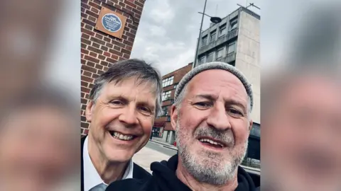 Two men pose for a selfie by a blue plaque which is mounted onto a wooden square on the side of a building.