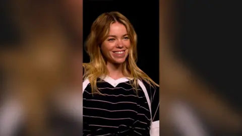 Milly Alcock has shoulder-length blonde hair. She is wearing a black top with thin horizontal white stripes.