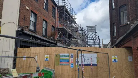 Wooden hoarding that's sealing off part of the main shopping street in Dorchester. Beyond the wooden panels is the collapsed building which is surrounded by scaffolding.