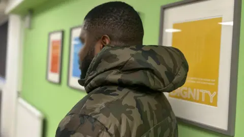 A man facing away from the camera with a khaki jacket. He has short hair. The room is green and there are frames on the wall. One frame has a yellow photo with the word 'Dignity'.