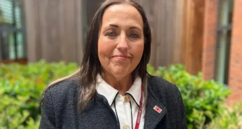 Jo Monk stands in front of foliage that is outside a brick and glass building. She has shoulder-length brown hair and is wearing a grey jacket and white blouse. She is smiling at the camera.