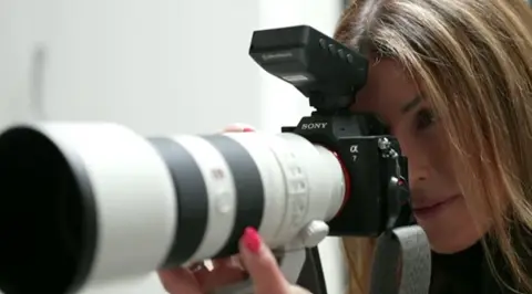 Jane Jordan uses a long lens camera. She is pictured holding it up to her face and using her left hand to steady the lens. She has long light brown hair. 