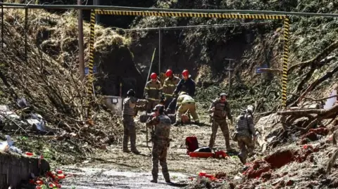 Uniformed officers search the site of the explosion in Quatro Barras, Brazil. Debris, including tree trunks and branches, can be seen scattered across the site. Some officers carry shovels.