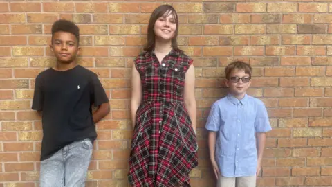 Simon Thake/BBC Two teenage boys and a teenage girl line up against a brick wall in their best clothes