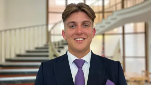George Finch, the Reform leader of Warwickshire County Council, stands in front of a spiral staircase. He is wearing a navy jacket, white shirt and purple tie.