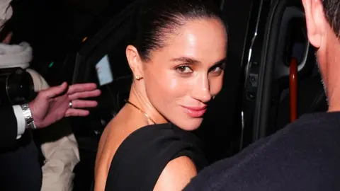 Close up of Meghan Markle at a red carpet event surrounded by security wearing a black evening dress, looking over her shoulder