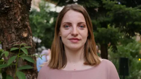 Majana Koceku is smiling directly at the camera. She has dark blonde hair in a long bob and is wearing pink top. Behind her are some trees. 