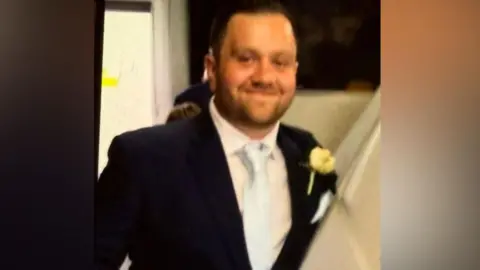 A man in a black suit with a light blue tie.