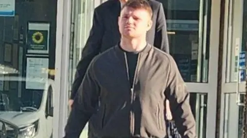 A man wearing dark clothing walking out of a building. The sun is shining on his face. He is looking the the left of the camera. A glass window is behind him and a car is in the reflection. A person is walking behind him. 