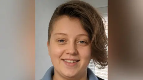 Family photo Menna, a young woman with short dark hair which is shaven at the sides, and has a long quiff at the front. It is a head and shoulders shot of her, with a window with blinds behind her. 