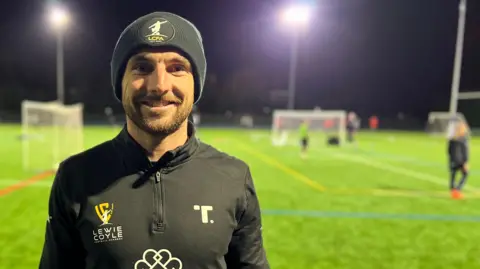 BBC Lewie Coyle, a professional footballer, stands on a floodlit football pitch as children play in the background. He is wearing a black beanie hat and a black sports top displaying the logo of his football academy.