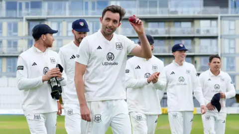 Glos seamer Matt Taylor holds the ball aloft in front of his team-mates after taking five wickets against Northants