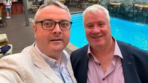 JUAN WATTERSON Juan Watterson, who has short grey hair and glasses, taking a selfie of himself and John Wannenburgh in front of a swimming pool.