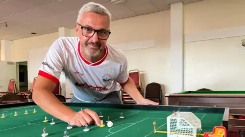 BBC Aaron Skinner wearing white t-shirt and leaning over Subbuteo table with miniature players on.