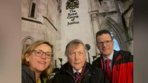 Aska Pickering Aska Pickering with long blonde hair and glasses, Andrew Stevenson with medium-length grey hair wearing a blue and red tie, and Simon Collyer with short dark hair and glasses wearing a red coat and red tie, in front of the Royal Courts of Justice - a gothic light stone building with a crest and "The Royal Courts of Justice" on one of the walls.