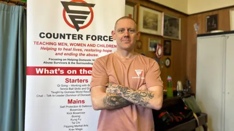 BBC News Scott in a pink t-shirt with a Counter Force logo, he's bald and has his arms crossed with tattoos on both arms. Standing in front of a Counter Force sign.