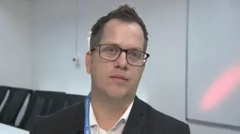 Mark Slemmings/BBC Clive Phillips sits in an office. He has dark-framed glasses and is wearing a white shirt and a dark suit jacket