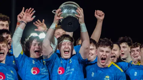 Ben Lumley/BUCS A close up of rugby players all looking ecstatic. A man in the middle holds up a silver trophy with his mouth wide open in joy.
