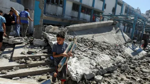 Reuters A Palestinian boy carries pieces of wood following an Israeli strike on a Unrwa-run school in Nuseirat refugee camp, in central Gaza (16 July 2024)