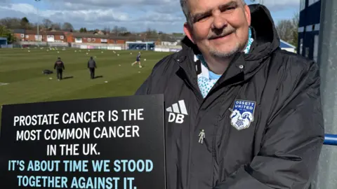 Prostate Cancer UK The image shows a scene at a football ground, positioned beside the pitch. The background includes the playing area where several individuals are visible, as well as buildings and trees surrounding the venue. A man is positioned on the right-hand side of the image, standing close to a barrier near the pitch. He is wearing a dark Ossett United jacket featuring the club’s crest and the initials “DB” on the chest and is holding a large signboard. The sign is black with white and blue text and reads: "Prostate cancer is the most common cancer in the UK. It's about time we stood together against it."