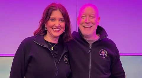 A woman with dark hair hugging a bald man man in a radio studio with a purple backdrop 