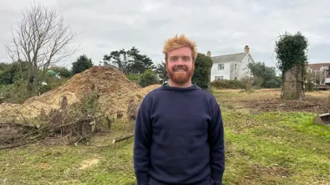 Jack Etheridge wearing a thick navy blue jumper. He is stood in front of farm land which includes a pile of woodchip, trees and houses in the background. 