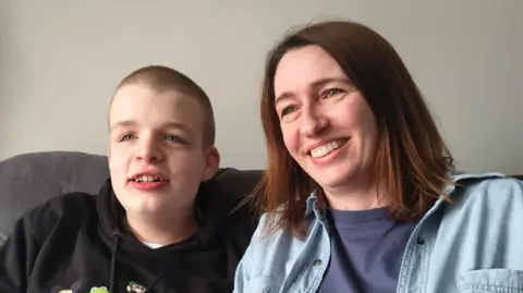 Teenager William sits on a sofa with his mum Sarah. William has a shaved head and wears a black hoodie. Sarah has short brown hair and wears a denim shirt with a navy T-shirt underneath. They are both smiling and looking away from the camera.