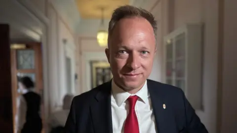 A man with brown greying hair stares into the camera. He has a black jacket, white shirt and red tie.