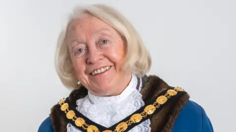 City of Wolverhampton Council A woman with blonde hair is wearing a chain and partly white and blue clothing. She is in front of a plain white background.