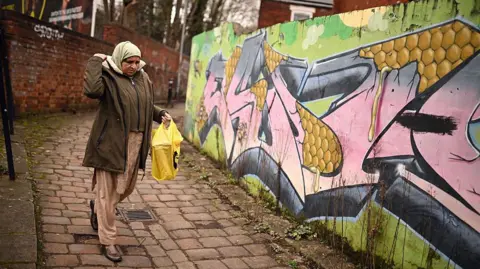 A woman passes graffiti sprayed on the wall of an alleyway in Longsight, within the Gorton and Denton parliamentary constituency