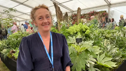 Clare Worden/BBC A woman wearing a navy dress stands in front of a square garden full of green and white leaves, flowers and stems. In the centre of the garden is a dead tree. 