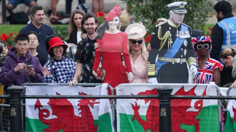 Reuters People in the crowd smile while holding cutouts of King Charles and the Princess of Wales behind a fence draped with two Welsh flags