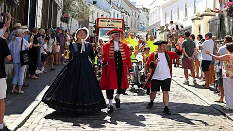 People walk through part of Alderney as part of a parade during Alderney Week. Two men leading the procession are dressed in red town crier style clothes. A woman is wearing a black Victorian dress. Crowds are stood either side of the road.