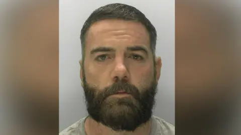 Police headshot of Robert Hall. He has a full beard and short brown hair, brown eyes and is wearing a grey t-shirt.