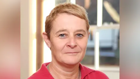 BTG Julie looks at the camera in a close up head shot. She has short cropped blonde hair and wears a gold necklace and red knitted top.