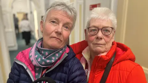 BBC A women with grey hair in a blue jacket wearing a scarf with pink hearts stands next to another woman with grey hair and glasses in a red jacket