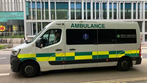 A white ambulance van captured from the left side, with hi-vis yellow and green decals along the bodywork and the word 'Ambulance' at the top on capitals. In the background you can see the Bristol Royal Infirmary building with large glass windows. 