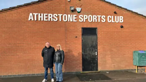 BBC The image shows a man in a knee-length black sports coat, standing next to a woman with shoulder-length blonde hair, who is wearing a black leather jacket over light blue jeans. They are both standing in front of a black door in a red brick wall, under a sign saying 'Atherstone Sports Club'.