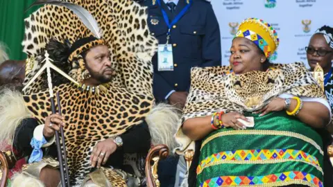 Getty Images South Africa's Zulu king and his archetypal  woman  are seen successful  colourful accepted   attire during an authoritative  event. The king is wearing a leopard people     wrapper  implicit    his achromatic  agelong  sleeved apical  portion    the queen is besides  wearing a akin  wrapper  implicit    her accepted   greenish  dress.