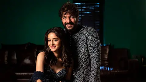 Hindustan Times/Getty Images Ananya Panday sitting in her home, with her father Chunky Panday standing next to her. They are wearing sparkling traditional Indian outfits and smiling. Behind them in the room is a brown leather sofa and green colored walls.