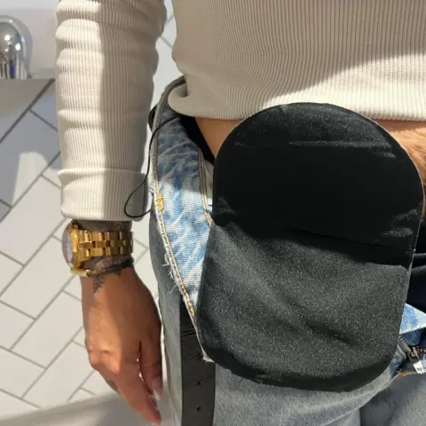 Close up photo of Jenna Robinson's stoma which is attached to her lower abdomen. She is wearing jeans and a white jumper. The stoma is a small bag which is often fitted because of bowel diseases and attach to a hole - or stoma - in the abdomen, allowing waste to be diverted out of the body.