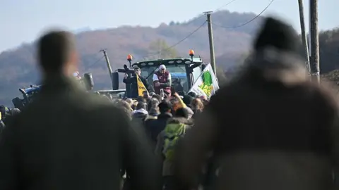 AFP via Getty Images President of the Rural Coordination in Ariège Sebastien Durand and regional figure of the agricultural protest movement Jerome Bayle wave flags and chant to a crowd, who are attending a farmers' protest against the slaughter of a 200-cow herd, following detection of lumpy skin disease (LSD) in Les-Bordes-sur-Arize, France. 