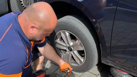 George Mircas George squats down beside a car wheel, holding some nuts and bolts in his hand. He wears orange gloves and wears a blue and orange T-shirt. He is not facing the camera. 