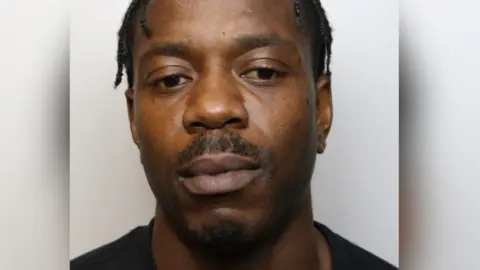 Richard, who has dark hair in short twists and short stubble on his face, looks down and away from the camera in a custody picture. 