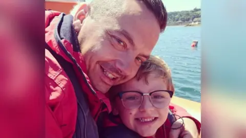 Dean and Charlie Lowe. On the left, father Dean has short dark hair, is wearing a red jacket and is smiling broadly, with his arm around Charlie. On the right, Charlie is wearing glasses, has light brown hair and is also smiling widely