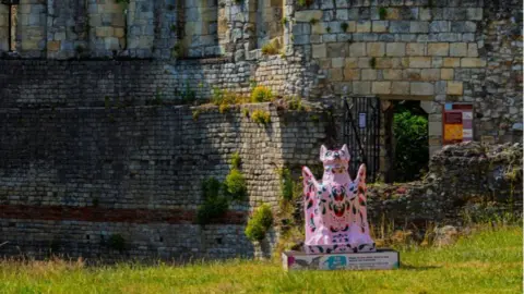A pink-coloured sculpture of a grotesque creature placed near ruined walls. It sits 