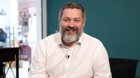 Charles Radclyffe in a medium close-up shot, sitting at a table with a teal-coloured wall across most of the background. He has a beard and his hair is dark and greying, he wears a white shirt and is smiling at the camera.